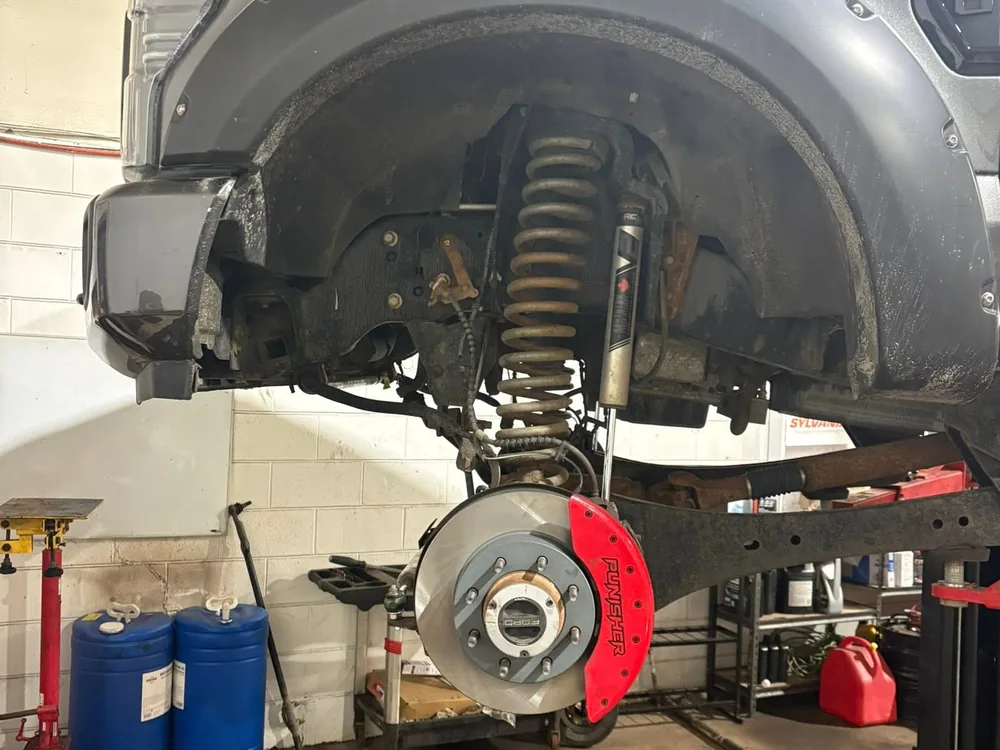Front suspension of a pickup truck raised on a hoist at AutoNorth Auto Repair in North Bay, showing the coil spring, shock absorber, brake rotor and a red brake caliper with the bumper removed