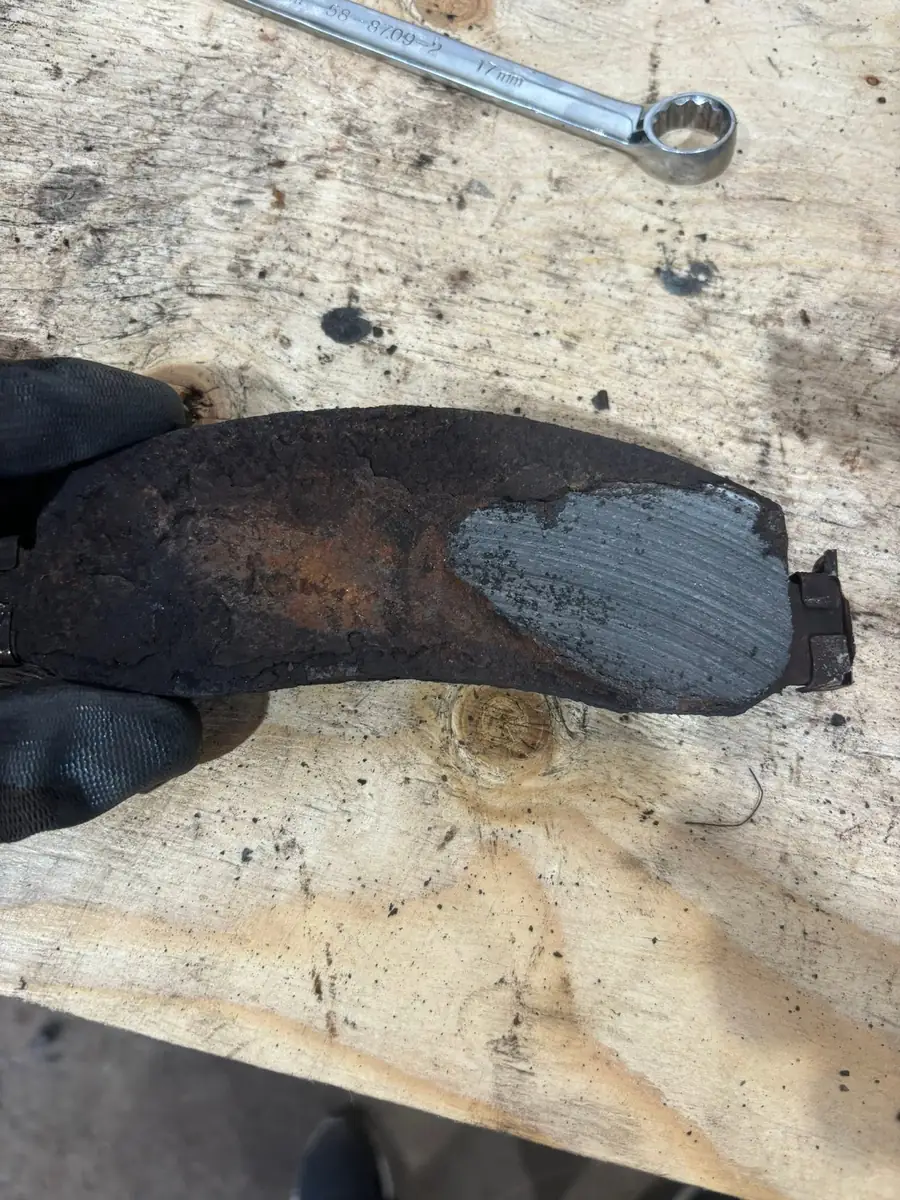 Gloved hand holding a rusted brake pad worn down to a thin strip of friction material on a wooden workbench beside a 17 mm wrench at AutoNorth Auto Repair in North Bay